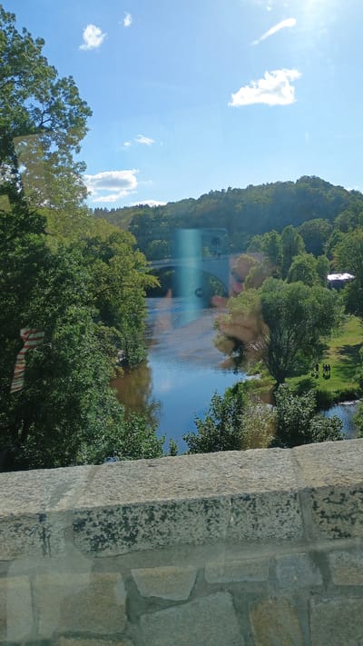 The image shows a wide river with a stone bridge spanning across it in the distance. The river is bordered by lush green trees on both sides, with a gentle hill covered in foliage forming the background. The sky is a vibrant blue with a few white clouds and the sun is visible, casting a bright glare on the water and sky. In the foreground, a stone wall made of large, irregular blocks is visible, suggesting it is part of a bridge or elevated structure. On the right bank of the river, a grassy area is visible with a small group of people gathered. The overall scene depicts a tranquil natural landscape with architectural elements.