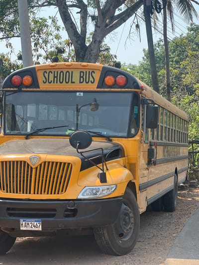 A yellow school bus is parked on a paved road in Santa Cruz de Yojoa, Honduras. The front of the bus, including the "SCHOOL BUS" signage, headlights, and front grille, are visible. The bus has a license plate that reads "HAM 2467" and indicates it is from Honduras, Central America. Trees and power lines are visible in the background.