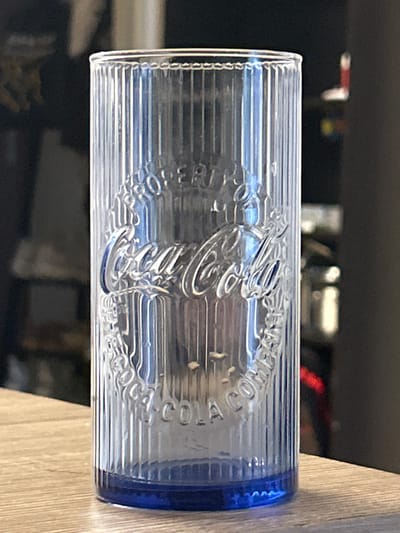 A clear, fluted glass with a blue base sits on a wooden surface. The glass has embossed lettering that reads "Coca-Cola" in the center, surrounded by a circular design. The background is blurred, showing a dark, indistinct environment with hints of what might be shelves or objects. The image is a close-up shot, focusing on the glass. The location is not ascertainable from the image.