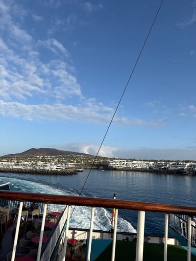 The scene depicts a view from a cruise ship sailing past Playa Blanca, Spain. The ship's railing and deck, with tables and chairs, are visible in the foreground. The water churns as the ship moves, creating whitecaps. In the distance, the shoreline of Playa Blanca is lined with white buildings. A mountain rises behind the town. The sky is blue with scattered clouds.
