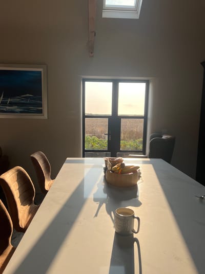 An interior shot depicts a sunlit room in Skipsea, United Kingdom, overlooking a rural landscape. A large, rectangular white dining table occupies the foreground and midground, reflecting strong sunlight that creates distinct, elongated shadows of objects. On the left side of the table, three upholstered beige dining chairs with fluted backs are visible. Centered on the table is a light wooden bowl containing bananas and other packaged items. To the right of the bowl, a white ceramic mug with an embossed pattern casts a long shadow.
The background features a set of dark-framed double doors, providing a view of an extensive golden-brown field, likely cultivated with crops such as wheat or barley, and a foreground patch of green leafy plants. The sky visible through the upper portion of the doors is bright and hazy. To the left, a framed painting depicting white sailboats on a dark blue sea is mounted on a light-colored wall. To the right of the doors, a dark grey upholstered armchair with visible studs is partially visible. The room has high ceilings, a prominent wooden beam, and a skylight overhead, admitting additional natural light. No individuals are present in the scene.