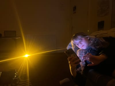 This image depicts an indoor scene, likely a bedroom, in a dimly lit environment, suggesting nighttime or a low-light setting. A young person, appearing female with light-colored hair and bangs, is positioned on the right side of the frame, reclining or lying down. They are engrossed in a smartphone, which emits a distinct blue light that illuminates their face. They are wearing what appears to be a denim jacket with a fluffy, possibly faux fur, collar or trim.
A very bright, yellow light source with prominent lens flares and streaks emanates from the lower-left side of the image, dominating that half of the frame. On a table in the mid-left, a rectangular electronic device, resembling a speaker (possibly Marshall branded), is visible with a cable connected. Below the table, a white, horizontally ribbed radiator is mounted on the wall.
In the background on the right, a closed, light-colored door is visible. Above the door and to its right, several documents or posters are affixed to the wall. One clearly visible sign is a blue and white "FIRE ACTION" notice. The room appears to have a carpeted floor. The overall atmosphere is one of quiet solitude or relaxation. This scene is located in a room in Dundee, United Kingdom.