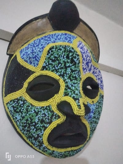 A black, ornate mask, heavily adorned with multi-colored beads, is depicted at a slight angle against a light-colored, possibly white or off-white, wall. The mask has a generally oval face with recessed, elongated oval eye openings, a prominent, sculpted nose, and full, sculpted lips forming the mouth. The base material of the mask appears to be dark, likely wood or a similar hard material.
The mask's surface is intricately covered with small, circular beads in distinct color sections. Yellow beads outline the facial features, including the eye sockets, the bridge and ala of the nose, and the contours of the lips. They also form curvilinear patterns across the forehead, cheeks, and chin. The areas between these yellow bead outlines are filled with clusters of other bead colors: vibrant blue beads are prominent on the upper right and left sides of the forehead, transitioning into lighter blue and green beads across the cheeks and lower face. Some sections, particularly on the far left side of the face, are covered in black beads. A rounded, black object, possibly a cap or hair bun, is affixed to the top of the mask, resting on a broader, gold-colored, crescent-shaped piece that forms part of the mask's upper structure.
The background is uniformly plain, suggesting an indoor display setting. No other individuals, objects, or environmental details are discernible to indicate a specific location within the city of Riabai ward, Kenya. A digital watermark "OPPO A53" is visible in the lower left corner of the image, indicating the capture device.