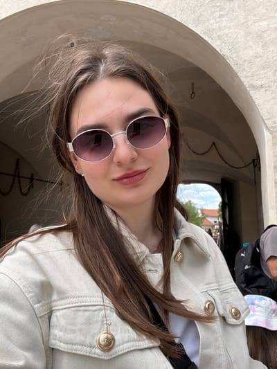 A young woman is posing for a picture inside a stone archway, presumably part of the Červený Kameň Castle in Častá, Slovakia. She is wearing sunglasses and a light-colored jacket with prominent gold buttons. In the background, through the archway, there is a glimpse of trees and building tops, suggesting an outdoor environment. There is a chain hanging in the archway. A person's head and a black bag are partially visible in the background, above the woman.
