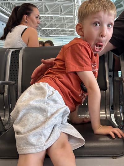 A young Caucasian boy with blond hair and wide green eyes is captured in a close-up, low-angle shot, sticking his tongue out at the camera. He is wearing a bright orange t-shirt and light grey shorts. He is seated on a dark grey airport bench, with his body leaning forward and his hands resting on the seat. To the left of the boy, a woman with dark brown hair in a ponytail, wearing a grey tank top, is visible from the shoulders up, looking away from the camera with a neutral expression. In the background, a blurred environment suggests an airport terminal with a high ceiling structure. The image appears to be taken in the city of Bol'shie Kabany, Russia.