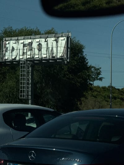 A billboard is covered in graffiti. The graffiti reads "DEION" in large, stylized white letters with black outlines. Above the large letters, smaller white text is visible, possibly an ad. The billboard is supported by metal poles and sits behind a dense green treeline. In the foreground, the roof of a dark-colored car is visible. The scene appears to be captured from another vehicle, as a portion of a windshield is in the upper frame. The sky is a clear, pale blue. The location is identified as Leganés, Spain.