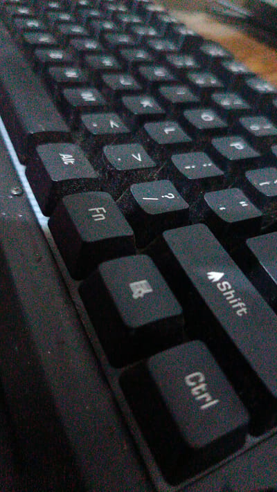 A close-up, grainy, slightly tilted, overhead shot of a black keyboard shows a portion of the keys. In the foreground, the "Ctrl" key is visible, followed by the "Shift" key with an upward arrow. To the left, the "Fn" key is prominent. Other keys with symbols like a question mark, comma, and period are also visible, as well as alphanumeric keys. The lighting is dim, casting shadows on the keys and creating a speckled appearance across the image. The background is dark and out of focus, suggesting an indoor setting. There is no information in the image to determine its location in Baubau, Indonesia.