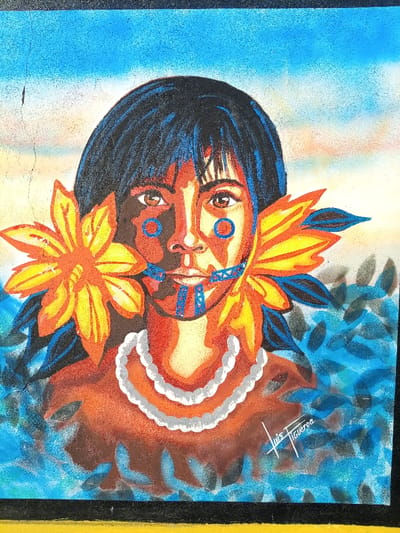 A vibrant mural in Cumaná, Venezuela, depicts a stylized portrait of an indigenous individual from the chest up. The subject has dark, shoulder-length hair and brown eyes. Distinctive blue geometric markings are present on the cheeks, resembling circles, and on the chin and mouth area, forming intricate patterns. The skin tone is rendered in shades of reddish-brown. Large, stylized floral or feather arrangements in yellow and orange protrude from each side of the head. A multi-strand white beaded necklace adorns the neck. The background transitions from a bright blue sky at the top to a pale, almost white horizon, with lower portions featuring blue-green, leaf-like foliage. The artist's signature, "Luis Figueroa," is visible in white on the lower right chest area of the figure. Minor vertical cracks are evident on the painted surface, particularly in the upper left section.