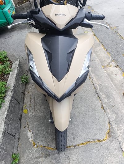 This is a frontal, eye-level medium shot of a beige and black Honda scooter parked on a concrete surface. The scooter's handlebars, front wheel, and a portion of its body are visible. The scooter has a sleek, modern design with sharp angles. The word "HONDA" is visible in white text on the front of the scooter's fairing.

The scooter is parked on a street next to a concrete curb. Green plants grow on the left side of the curb, and yellow petals are scattered on the ground and in the cracks of the concrete, suggesting it might be daytime and a day with some wind or a tree shedding its flowers. The ground is made of cracked concrete, indicating an urban or semi-urban environment.

To the left of the scooter, a portion of a green vehicle is visible, possibly another scooter or a small car. The background is slightly blurred, but it appears to be an outdoor setting with a road surface. The lighting suggests it is daytime, and the weather appears to be overcast or bright but not direct sunlight. There are no people visible in the image. The scene is static, with the scooter parked and stationary. The overall impression is one of a typical street scene in a location like Imus, Philippines, with a focus on the parked scooter.