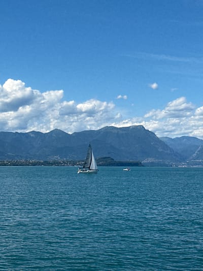 This is a vertical, eye-level, outdoor, wide-angle photograph of Lake Garda, Italy, from the perspective of the city of Desenzano del Garda. In the foreground is the aquamarine water of the lake, with small ripples on its surface. In the middle ground, a white sailboat with black trim and a single white sail is sailing from left to right across the lake. The sailboat has two visible figures on board. Further to the right in the middle ground, a small white motorboat with two visible figures is moving from left to right. In the background are the blue-green mountains and hills of the opposite shore of the lake, with a scattering of buildings visible on the lower slopes. The sky is a bright blue, with scattered white cumulus clouds.
