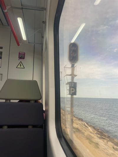 The image shows the interior of a train carriage looking out a window. Inside the carriage, there are seats and overhead lighting. Outside the window, there is a view of the Mediterranean Sea, with a rocky shoreline. A railway signal, marked with "32" and a "P" symbol, is visible along the track. The sky is cloudy. The train appears to be traveling along the coast of Mataró, Spain.