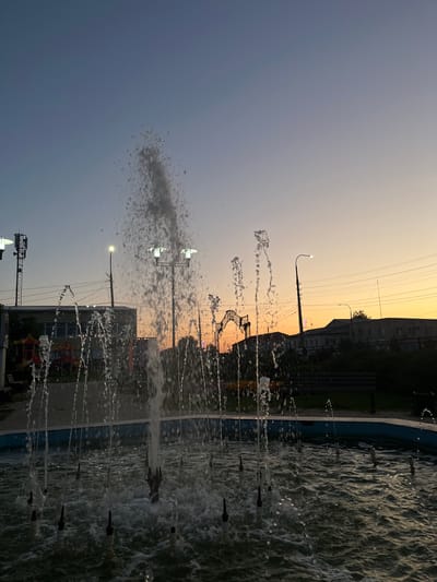 A fountain with multiple jets of water is in the foreground, set within a circular basin. The water is illuminated by the twilight sky, casting a shimmering effect on the surface. In the background, a street scene unfolds at dusk. Buildings and streetlights are visible, along with power lines crisscrossing the sky. A playground structure with colorful elements is situated to the left. An arched gateway is visible further in the distance. The overall setting appears to be a public park or plaza within the city of Preobragenskaya, Russia, as indicated by the surrounding urban infrastructure and the presence of recreational facilities. The sky displays a gradient of colors, transitioning from a deep blue at the top to a warm orange and yellow near the horizon, suggesting the sun has recently set. There are no people visible in the image.