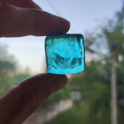 A close-up, eye-level shot shows a person's thumb and index finger holding a small, translucent, blue, cube-shaped object. The object appears to be made of glass or resin and contains small air bubbles and internal striations. The background is blurred, showing out-of-focus trees and a hazy sky, suggesting an outdoor setting. The image does not provide any specific clues about the location in Kineshma, Russia, or any details about the person beyond their fingers.