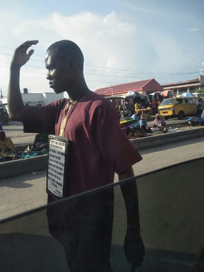 A dark-skinned adult male, identified as Adamu by a sign, is depicted in profile from the waist up, facing right. He wears a maroon-colored short-sleeved top with gold-colored embroidery around the neck. A white rectangular sign is suspended from his neck by a thin strap, displaying black text that reads, "TO WHOM IT MAY CONCERN / MAY GOD BLESS YOU / AM ADAMU, I AM DUMB / AND DEAF, PLEASE / PLEASE HELP ME FOR HALL / GMENTS TO BETTER THAN / NG MAY THE ALMIGHTY / GOD HELP YOU IN ALL / OUR UNDERTAKINGS / (AMEN) / THANKS / PLEASE HELP ME." His right arm is raised, with the hand open, positioned near his forehead as if shielding his eyes from sunlight. The image is captured from within a vehicle, with a dark window edge visible across the lower foreground.
The background reveals a bustling outdoor market street scene in Awjaw, Nigeria, under a bright, cloudy daylight sky. People are visible engaged in market activities, some seated on the ground along a curb. Market stalls with various goods are present, and multiple umbrellas provide shade. A yellow commercial passenger van is parked or moving on the right side of the street. Buildings with red-tiled roofs are visible in the mid-ground, and lighter-colored buildings with a dark structure on top are visible further left. A slender, tall structure, possibly a minaret, is faintly visible on the far left horizon.