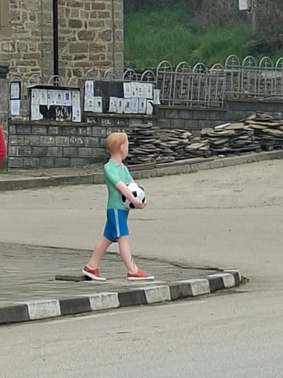 A color photograph depicts a statue of a child holding a soccer ball in Debnevo, Bulgaria. The child is wearing a green shirt, blue shorts, and red shoes. The statue is positioned near a curb painted with alternating black and white stripes. Adjacent to the walkway and statue is a building of cut stone and a fence that houses various publications. A grassy area is located to the right of the fence.