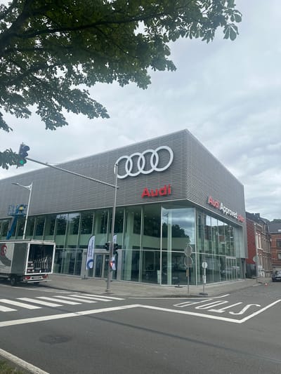 This media file captures an exterior view of an Audi dealership in Liège, Belgium. The building features a modern architectural design with a facade covered in a patterned metallic material and large glass windows. Prominently displayed on the building are the Audi rings logo and the text "Audi" and "Audi Approved plus" in red lettering. The scene is a street in front of the dealership, with a road, a pedestrian crossing, and a sidewalk. A traffic light shows a green signal, indicating traffic is allowed to proceed. A stop sign is painted on the road, advising vehicles to halt. A white delivery truck with its back open is parked near the entrance. In the foreground, a large tree with green leaves partially obscures the view of the building and the sky. The sky is overcast, suggesting cloudy weather. The overall impression is that of a bright, albeit cloudy, day, with the dealership serving as the central focus of the scene.