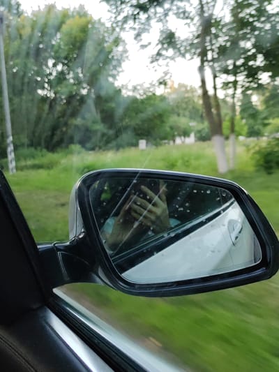 The image shows a reflection of a person's hands holding a smartphone in a car's side mirror. The mirror is slightly dirty with water droplets. The car is in motion, indicated by the blurred green landscape and trees visible through the car window and in the distance. The scene suggests the person is a passenger or driver, capturing a moment while traveling through a rural area. The location is identified as the Lyshchytsy Village Council, Belarus.