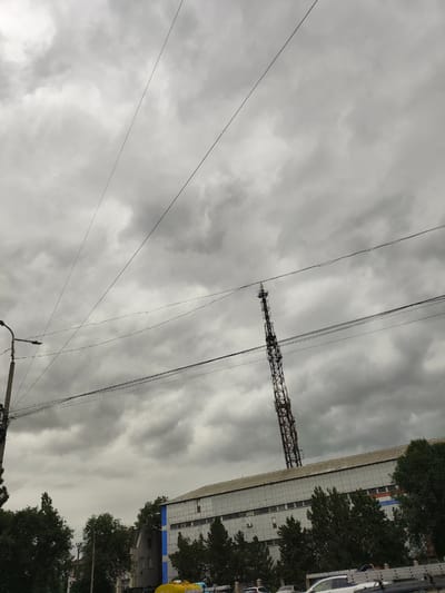 The image shows an overcast sky with thick, gray clouds. Several wires are strung across the frame, intersecting at various angles. In the middle ground, a tall, lattice-like tower, possibly a radio or telecommunications tower, is visible. Below the tower, a long, white building with many windows and a metallic roof stretches across the scene. Trees with dark green foliage are present in front of the building. On the left side of the image, a lamppost with an attached light fixture stands. A portion of a blue and white building is also visible amongst the trees. The scene appears to be in an urban or industrial area.