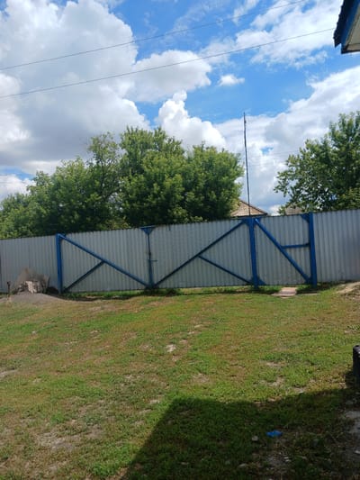 The image shows a galvanized metal fence with blue trim and a double gate, located in the city of Жалтыр, Kazakhstan. Behind the fence, there is a line of green trees and a section of a house with a brown roof. The sky above is partly cloudy with a bright blue sky peeking through. In the foreground, there is a grassy area with some dirt patches and a shadow cast from the right side of the frame. To the left of the gate, there are some concrete blocks and a tree stump. The overall scene depicts a residential or semi-rural outdoor setting.