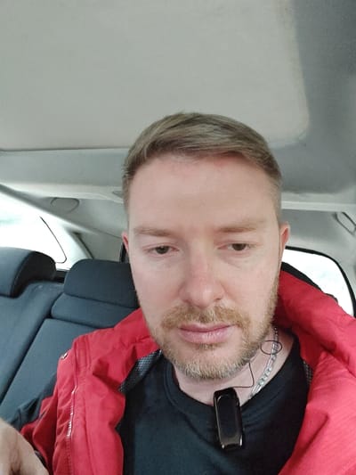 A male subject with light brown hair and a short, light brown beard is depicted in a close-up, seated inside a vehicle. He is wearing a black crew-neck shirt beneath a red, possibly insulated, jacket. A silver-colored chain is visible around his neck, from which a small, black, glossy rectangular electronic device hangs. His gaze is directed downwards. The vehicle's interior features a light-colored headliner and dark grey or black rear passenger seat headrests. A rear window is visible, showing a bright, undifferentiated outdoor environment. The specific location within the city of Nesher, Israel, cannot be determined from the visual information present in the image.