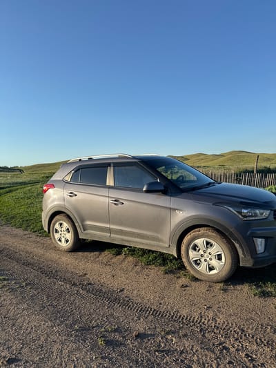 A grey SUV is parked on a dirt road in Starokhalilovo, Russia. The car is viewed from the side, with its front right tire on the road and its rear right tire on grass. The car has tinted windows and a roof rack. In the background, there is a wooden fence and rolling green hills. The sky is clear and blue, indicating a sunny day. The image appears to have been taken in the late afternoon or early evening, as suggested by the warm lighting and long shadows. There is no text visible in the image.