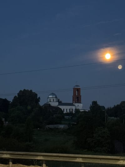 The image depicts a twilight scene in Svatkovo, Russia, featuring a church complex surrounded by trees. Two celestial bodies, likely the moon and a smaller satellite or planet, are visible in the deep blue sky, with one illuminated by the setting sun and the other faintly visible. A cluster of buildings, including a church with a silver dome and a brick bell tower, is nestled among the trees in the mid-ground. The foreground is dominated by a wooden barrier, suggesting the vantage point is from a road or elevated area. The overall atmosphere is calm and serene, with the fading light casting long shadows and creating a sense of depth.