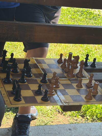 The image shows a close-up view of a chessboard with black and brown pieces. The chessboard is placed on a wooden surface, possibly a table or bench, outdoors. Green grass and sunlight are visible in the background, suggesting a park or garden setting. Two individuals, partially visible, are seated on either side of the chessboard, engaged in a game of chess. One person is wearing black shorts and a blue shirt, while the other is wearing black shorts and a grey t-shirt. Their legs and feet are visible, wearing athletic shoes and socks. The game appears to be in progress, with several pieces already moved on the board. The chessboard itself is made of cardboard or a similar material and has numerical and alphabetical markings on its edges. The scene depicts a casual outdoor activity, likely in Dinkata, Bulgaria, given the prompt's contextual information.