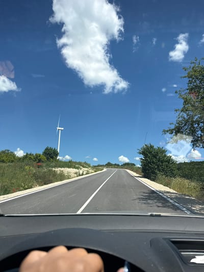 The image depicts a road winding through a rural landscape under a clear blue sky dotted with white clouds. A single white wind turbine stands tall in the distance to the left of the road, with another smaller one further back. The road is paved and has white lane markings. The sides of the road are lined with green grass, some wildflowers, and scattered trees and bushes. The foreground shows a portion of a car's dashboard and a hand on the steering wheel, indicating the image was taken from inside a moving vehicle. The environment suggests a sunny day in a somewhat hilly or mountainous area. The scene is consistent with the landscape found in the vicinity of Suvorovo, Bulgaria, known for its wind farms and rolling terrain.