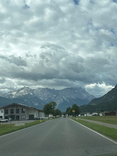 The image depicts a road leading towards the city of Farchant, Germany, with a backdrop of the Wetterstein Mountains. The sky is overcast with heavy, grey clouds. On the left side of the road, a modern, multi-story building with "KFZ" signage is visible. Several cars are parked or driving on the road and in front of the building. Green grass and trees line the road, with a few houses and other buildings scattered in the distance on the right side. The road appears to be a two-lane highway with a white line marking the center. The overall scene conveys a somewhat cloudy and cool atmosphere.