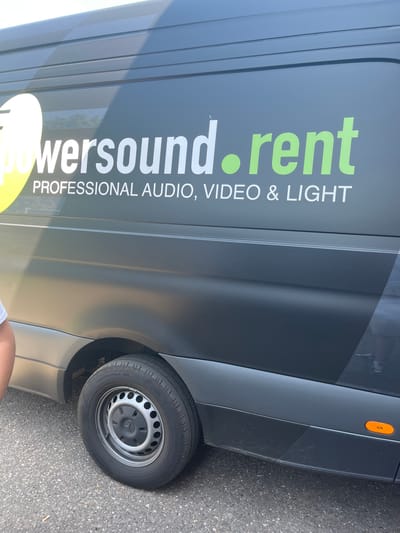 The image shows the side of a dark gray van with text and graphics on its side. The text reads "powersound.rent" in large white letters, with "powersound" having a yellow oval logo integrated into its design. Below "powersound.rent," in smaller white letters, is "PROFESSIONAL AUDIO, VIDEO & LIGHT."
The van appears to be parked on an asphalt surface, possibly a street or parking lot. The lighting suggests it is daytime, and the shadows are not particularly long, indicating it might be mid-morning or afternoon. There are no strong indicators of specific weather conditions, but the surface is dry. The background is slightly out of focus but shows the typical environment of a town or suburban area, with some hints of greenery and buildings.
A portion of the van's rear wheel and fender are visible on the right side of the image. An orange reflector is also visible on the upper rear corner of the van. The overall scene is static, showing the van as the primary subject. There is a small portion of a person's leg visible at the bottom of the frame, suggesting a person is near the van, but their face or full body is not shown. The image is oriented vertically.