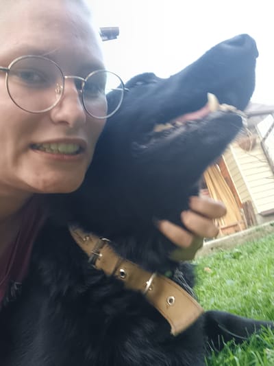A close-up, first-person perspective selfie features a person and a black dog outdoors. The person, wearing round, wire-rimmed glasses, is on the left side of the frame, with their face partially visible. They are smiling, revealing teeth. A black dog occupies the right and central portion of the image, with its head turned towards the camera and its mouth open, showing its teeth and tongue. The dog is wearing a tan collar. The background suggests an outdoor setting with green grass and a light-colored building with windows visible. The location is specified as Koledino, Russia.