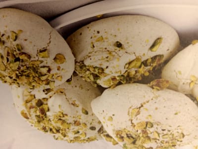 The image shows a close-up of several light-colored, irregular-shaped cookies, possibly meringues, dusted with chopped pistachios. The cookies are clustered together, with some overlapping. They appear to be on a white surface, possibly a plate or platter, with a textured gray background behind them. The lighting is soft, highlighting the textures of the cookies and pistachios. There are no people or animals visible in the image. The scene is a still life of food. The location in Rumaithiya, Kuwait, is not discernible from the image itself.
