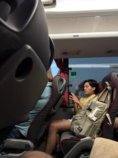The image shows the interior of a bus, likely in the city of Somma Lombardo, Italy, with a focus on passengers and seating. In the foreground, a dark gray bus seatback is prominently visible, partially obscuring the view of other passengers. To the right of this seat, a man is seated and looking at his phone. He is wearing a yellow and white striped shirt and light-colored shorts. A beige backpack with a white object attached to it rests next to him on the seat. Another passenger, with brown hair, is visible in the middle ground, wearing a blue and white striped shirt and dark pants. The bus interior features overhead storage compartments, air vents, and a window showing a blurred outdoor scene. Red fabric curtains are visible near the window. A sign with a green and white icon is attached to the wall behind the man. Seat numbers "45" and "46" are visible on a panel above the seats.