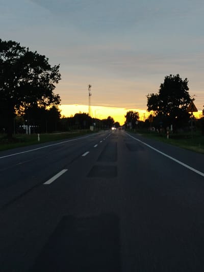 The image depicts a view down a rural road in Skierdy, Poland, at sunset. The road is asphalt and has white lane markings. On either side of the road, there are trees and vegetation. In the distance, the sky is a blend of orange, yellow, and blue, indicating the sun is setting. There is a cell tower visible on the left side of the road. There are also some road signs and streetlights visible. The image appears to have been taken from a moving vehicle.