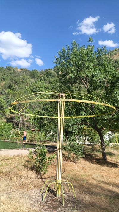 An outdoor, eye-level, medium shot captures a weathered, yellow metal merry-go-round structure in the foreground. The merry-go-round is comprised of a circular frame supported by vertical poles, with a spider-like base anchoring it to the ground. Dried, brown grass surrounds the structure, and a few small green bushes are visible. In the background, a swimming pool or pond with clear green water is situated to the left. A young man wearing blue shorts stands at the edge of the pool, near a yellow metal ladder leading into the water. Further in the background, a lush, tree-covered hillside rises under a bright blue sky dotted with fluffy white clouds. The overall scene suggests a recreational area, possibly abandoned or in disrepair, set within a natural, hilly landscape. The location is identified as Mirat, Tajikistan.