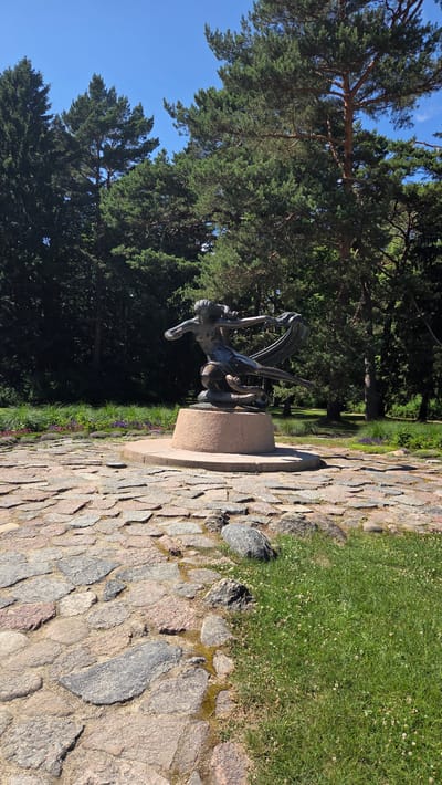 The image depicts an outdoor scene, likely in a park or garden, with a bronze sculpture as the central subject. The sculpture, which appears to be a female figure with drapery or wings, is mounted on a stone pedestal. The surrounding environment features lush green grass, a cobblestone path made of large, irregular stones, and tall evergreen trees. In the background, more trees create a dense, dark backdrop against a bright blue sky. The lighting suggests it is daytime, with clear weather. The overall impression is one of a serene, well-maintained outdoor space, possibly a memorial or a decorative feature in a botanical garden. The angle of the photo is from the side, capturing the sculpture and its surroundings in a somewhat tilted perspective. There is no visible text in the image.
