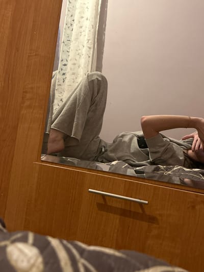 A person is reflected in a mirror attached to a wooden closet or cabinet. The individual wears a gray hooded sweatshirt and is positioned partially in the frame, facing the mirror. The mirror reflects a white floral patterned curtain and a blank wall behind the subject. The frame around the mirror is wooden and matches the cabinet. A silver handle is on the front of the cabinet, casting a shadow.
