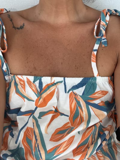 The image shows a close-up of a person's upper chest and shoulders, wearing a white top with a tropical leaf pattern in orange, teal, and blue. A small tattoo of a sprig is visible on the left shoulder. The skin appears tanned and has freckles. The background is a plain, textured surface, likely a wall or fabric. The image is cropped tightly on the subject and does not provide any context about the location within the city of Khashuri, Georgia.