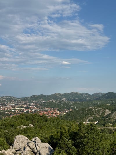 The image depicts a wide aerial view of the city of Dubovik, Montenegro, set against a backdrop of rolling green hills and a bright blue sky dotted with white clouds. In the foreground, large, grey, weathered rocks are prominent, suggesting a rocky terrain. Beyond the rocks, dense green trees cover the landscape, leading the eye towards the city.
Dubovik itself is visible in the mid-ground, characterized by a collection of buildings with red-tiled roofs, interspersed with other structures and some lighter-colored buildings. The city appears to be nestled within a valley or surrounded by the undulating hills. The hills themselves are covered in greenery, with patches of exposed grey rock or stone appearing in places. The overall impression is one of a rural or semi-rural settlement in a mountainous region. The sky is clear with scattered cumulus clouds, indicating fair weather. There are no visible people or animals in the image, and no specific actions or interactions are depicted. The scene is a static landscape view.