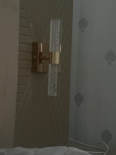 A bedside wall sconce with two cylindrical glass shades containing bubbles is mounted on a wall panel. The sconce features a brass-colored metal fixture. The wall behind the sconce is decorated with a subtle damask-style wallpaper. A white electrical cord is visible on the floor in the lower right corner of the image. The location is a room in Lebedinovka, Kyrgyzstan.