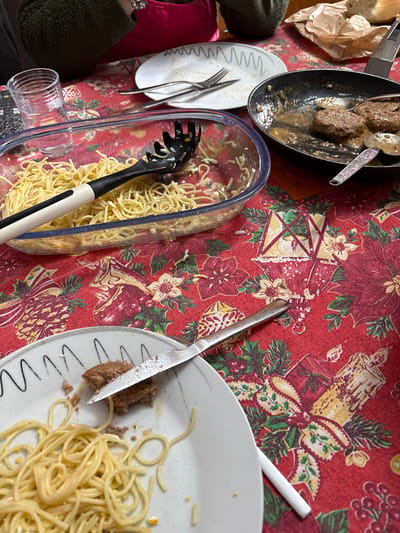 A table is depicted from a high-angle perspective, set with a red tablecloth featuring a festive pattern of green holly, red poinsettias, gold lanterns, and candles, indicative of a holiday season. The scene is located in Santa Maria da Feira, Portugal.

The table is laden with remnants of a meal. A large clear glass rectangular baking dish occupies the center-left, containing a substantial amount of yellow spaghetti with a black and white spaghetti server resting inside. To its right, a black frying pan holds three brown, circular meat patties submerged in a light brown gravy, with a silver spoon partially immersed.

In the lower foreground, a white plate with a black wavy rim contains a small portion of spaghetti and a partially consumed brown meat patty. A silver table knife rests across this plate. Further back and to the left, another white plate with a similar wavy rim is mostly empty, holding a silver fork and a silver spoon. A clear glass tumbler containing water is positioned behind the spaghetti dish.

In the upper-left background, the left arm and shoulder of an individual, dressed in a dark green or brown textured garment, are visible. A flash of magenta fabric is also visible, suggesting another person or a vibrant accessory. A crumpled brown paper bag, possibly containing bread, is visible in the upper-right background. The overall scene suggests a family meal, recently consumed or ongoing, with dishes still on the table.