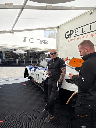The scene takes place outside a building in Stavelot, Belgium, at the coordinates 50.441, 5.969. Two men stand next to a white race car with orange accents and a black, blue, and white livery. The car is partially obscured by a white tent structure. The man on the left wears sunglasses, a black t-shirt with a logo on the left sleeve, and black pants. He holds a phone in his hand and looks at the man on his right. The man on the right wears a black sweatshirt with orange accents and a white "CODE" logo on his right sleeve. He holds a phone in his hand and looks at the ground. The car is numbered 25 and displays a logo for "SenseIT" and "Real Estate & Technology." There are red and white markings on the black, checkered, rubber-tiled ground. The building in the background has a black awning with a white sign that reads "GPELITE." The sign is upside down. The side of the building is painted white with the words "GPELITE DRIVE TO PERFECTION" on it. The words are reflected upside down on the roof of the tent. The background is partly obscured by the tent structure. The scene appears to be taken on a sunny day.
