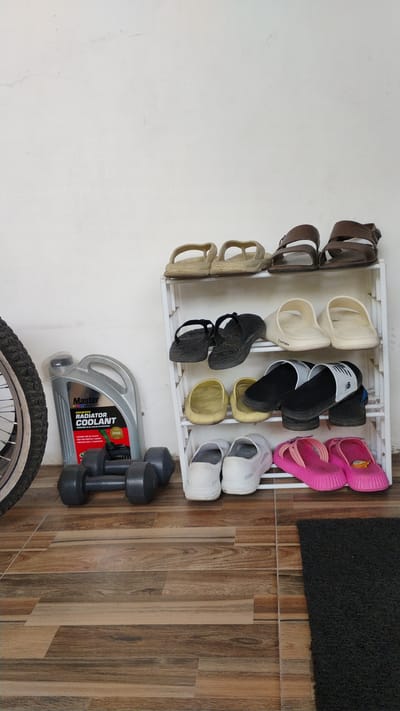 In Banjarsari, Indonesia, an outdoor scene unfolds against a pale, textured wall. A white, multi-tiered shoe rack is laden with footwear. The top shelf holds two pairs of light-colored sandals, followed by two pairs of black flip-flops on the second shelf. The third shelf displays two pairs of yellow clogs, and the fourth shelf presents two pairs of black slip-on shoes, one featuring a white "NB" logo. The bottom shelf showcases two pairs of white slip-on shoes and two pairs of bright pink flip-flops. To the left of the shoe rack, a grey jug of "Master Radiator Coolant" stands upright. Adjacent to the coolant jug are two dark grey dumbbells. A portion of a bicycle wheel with a black tire is visible on the far left. The ground is covered with wooden-plank-patterned tiles, and a dark grey rectangular mat lies on the right.