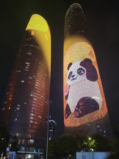 This media file captures a nighttime view of two cylindrical buildings in the Wuhou District of China, illuminated with a vibrant light display. The upper building prominently features a large, stylized image of a panda, with its face and body illuminated in white and black. The surrounding surface of the building is covered in a geometric pattern of warm yellow and orange lights, with scattered small, colorful dots that appear to be twinkling lights. The lower building is also covered in a similar geometric pattern, primarily in warm orange and yellow hues, with many small, brightly lit windows visible within the structure. The reflection of the upper building's light display is visible on the surface of the lower building, creating a mirrored effect. The scene is dark, indicating it is nighttime. There are no people clearly visible, but the illuminated windows suggest occupancy. Trees and other structures are partially visible on the right side of the frame, suggesting an urban setting. The overall impression is one of a festive and visually striking urban display.