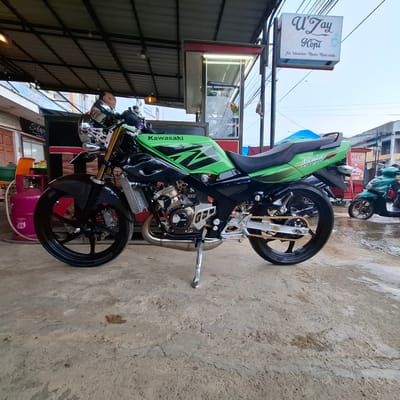 A green and black Kawasaki Ninja motorcycle is parked on a wet concrete surface under a covered area, possibly a roadside stall or shop in Rhieng Blang, Indonesia. The motorcycle has a sporty design with prominent green graphics on the fuel tank and rear fender, and carbon fiber accents. The front wheel, brake caliper, and suspension are visible. A pink gas cylinder with "135" marked on it is positioned to the left of the motorcycle, near a building entrance. In the background, a sign for "U'Zay Kopi" is displayed, along with other shopfronts and a green scooter. The ground is wet and reflective, suggesting recent rain. The overall scene depicts a common urban or semi-urban environment in Indonesia, with elements of commerce and transportation. There are no clearly identifiable people in the foreground, though a person's head is visible through the window of a small kiosk-like structure behind the motorcycle. The lighting suggests it is daytime, with overcast conditions.