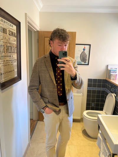 A male individual is visible in a mirror selfie taken within a brightly lit bathroom environment. The subject is a young adult male with light brown, curly hair, exhibiting a mullet-style cut. He wears a black long-sleeved shirt, a dark red tie adorned with yellow bee-like motifs, a brown and grey tweed-patterned blazer, light-colored trousers, and a brown leather belt. Accessories include a watch with a dark face and brown leather strap on his left wrist, a silver-colored ring on his left ring finger, and a silver-colored bracelet on his right wrist. His left hand is placed in his trouser pocket, while his right hand holds a green-cased smartphone to capture his reflection.
The background elements include a white ceramic toilet with an open lid on the right side of the frame, positioned against a wall tiled with black square tiles and a horizontal light-colored border. A white vertical radiator is situated between the subject and the toilet. A white ceramic sink with a white folded towel hanging beneath it is partially visible in the bottom right. The floor is covered with light-colored, possibly beige, rectangular tiles. On the left wall, a large, dark-framed print resembling a vintage newspaper advertisement with inverted text is displayed. On the back wall, to the right of a wooden door, a framed piece of art depicting a dark, textured object on a light background with blue and red accents is mounted. Several items, possibly bottles or dispensers, are visible on a shelf above the toilet. The image does not contain any visual indicators to confirm a specific location within the city of Hornsea, United Kingdom.