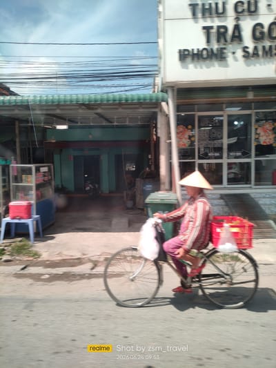 A blurred, daytime, outdoor scene captures a person riding a bicycle along a paved road in Ben Cau, Vietnam. The rider, wearing a conical hat, a red and white striped shirt, and purple pants, is carrying a red plastic crate filled with white plastic bags on the back of the bicycle. The bicycle has two wheels and appears to be in motion, indicated by motion blur.

To the right of the road is a white building with large glass windows, featuring signage with Vietnamese text, including "THU CU", "TRÀ GÓ", and "IPHONE - SAM". Some of the windows display images that appear to be advertisements. A dark green trash bin is visible in front of this building.

To the left of the road, a covered walkway with a green corrugated roof extends from a building. In front of this walkway, a glass display case containing various items sits on a blue base. A red cooler box rests on a small blue stool next to the display case. A motorcycle is parked in the entryway of a turquoise building behind the walkway. The overall environment suggests a commercial or residential area.