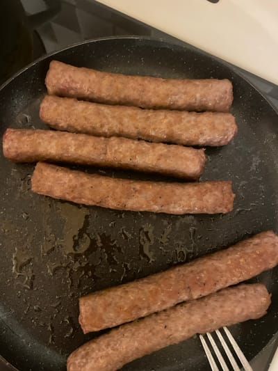 This image depicts a cooking scene in the city of Palež, Montenegro. Seven elongated, cylindrical meat items, consistent in appearance with sausages or ćevapi, are contained within a black, speckled non-stick frying pan. Five of these items are arranged horizontally in the upper-left portion of the pan, parallel to each other. The remaining two items are positioned diagonally in the lower-right section. The meat items exhibit a browned, grayish-pink exterior with visible searing marks. The pan's surface shows various droplets and streaks of cooking oil or grease. A silver-colored, four-tined fork is partially visible in the lower-right, its tines in contact with one of the diagonally positioned meat items. The frying pan rests on a dark stovetop, with a lighter, indistinct surface visible in the background.