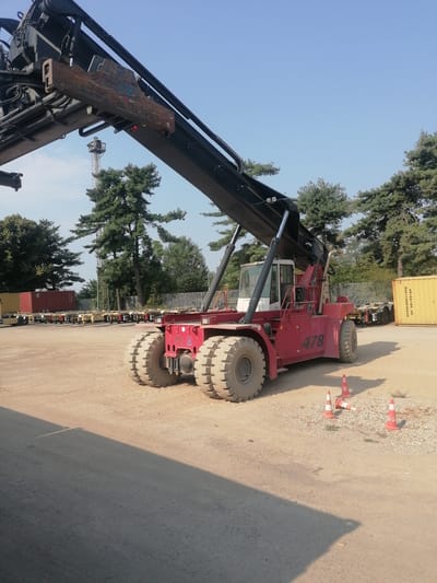 This image captures a bright, outdoor scene at what appears to be a shipping or logistics yard, possibly in Santo Stefano Ticino, Italy, given the context. The focal point is a large, red reach stacker, a heavy-duty vehicle used for lifting and moving shipping containers. The machine is positioned on a dirt or gravel surface. Its massive tires are clearly visible, as are its extended lifting forks at the top. The number "478" is prominently displayed on the side of the vehicle.
In the background, several shipping containers are lined up, some red and some yellow. A light-colored fence runs behind the containers, and beyond that, a line of trees, including pine trees, is visible against a clear blue sky. A tall, slender tower structure, possibly a communication tower or part of a crane, is also visible in the distance. There are three orange traffic cones on the ground near the reach stacker, one of which is tipped over. No people are visible in the image. The lighting suggests it is daytime, with shadows indicating the sun is at an angle. The overall environment appears to be an operational industrial or transportation hub.