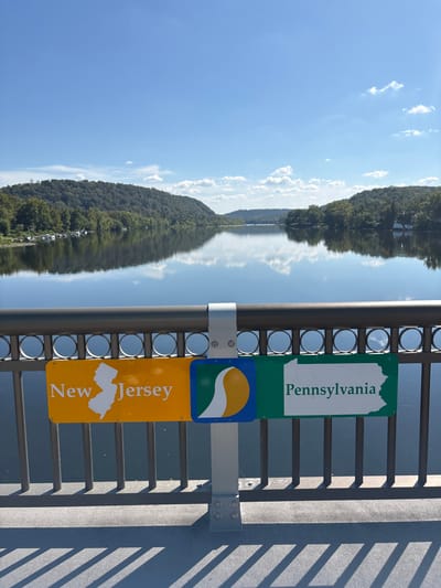 The image displays a daytime view from the Lambertville-New Hope Bridge over the Delaware River, separating Lambertville, New Jersey, from New Hope, Pennsylvania, under a clear blue sky with scattered white cumulus clouds.
In the immediate foreground, a metallic gray bridge railing, featuring horizontal bars and circular cutouts, spans the lower third of the frame. Attached to this railing is a vertical support bracket holding two prominent rectangular signs. The sign on the left is yellow and displays "New Jersey" alongside a white outline of the state. The sign on the right is green and displays "Pennsylvania" with a white outline of that state. Between these state signs is a smaller blue square sign featuring a white and yellow stylized "S" or wave motif. Sharp, parallel shadows from the railing structure are cast onto the light gray concrete surface of the bridge deck.
The midground is dominated by the calm, reflective surface of the Delaware River, mirroring the sky and distant landscape. On the left riverbank, a densely wooded area is visible, with several small, light-colored boats moored along the shore. On the right riverbank, also heavily forested, a few white residential-style structures are discernible nestled among the trees. Both banks feature rolling hills covered in lush green deciduous trees. The distant background consists of more tree-covered hills receding into the horizon beneath the expanse of the blue sky. No individuals are present in the scene.